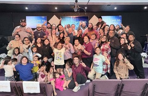 The Hosanna congregation, from little children to grandparents, smiling and holding up peace signs together in front of the stage, beneath screens that read Welcome to Hosanna.